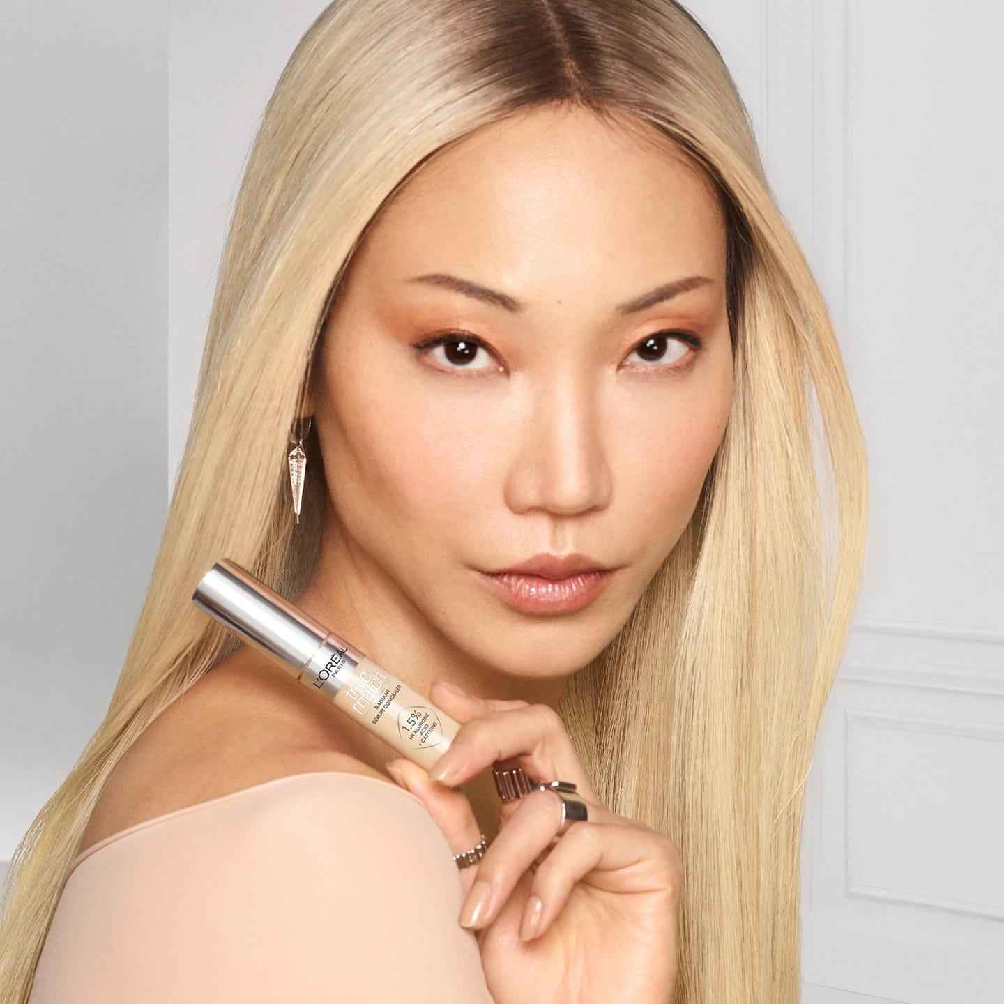 Eine junge Frau mit langen blond gefärbten Haaren schaut zur Seite und hält den Concealer 2R Light Medium in ihrer Hand